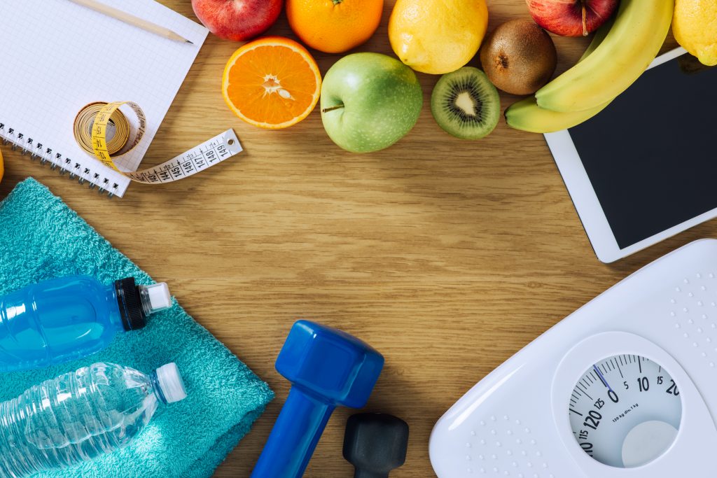 Weight loss concept with scale, dumbbell, healthy drink, fruit, towel, measuring tape and tablet on a table.