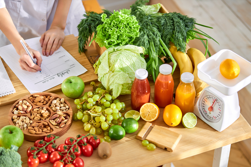 A table with colorful healthy foods, including fruits, vegetables, nuts, and natural juices, while someone writes a meal plan preparing balanced meals.