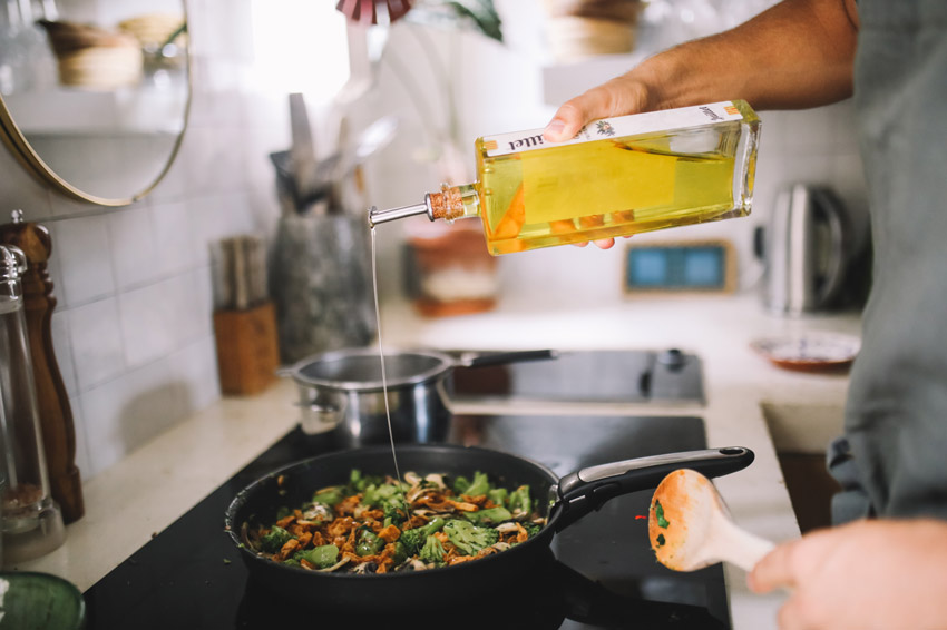 A man cooking a sautéed vegetable dish on an electric stovetop, pouring oil into a pan