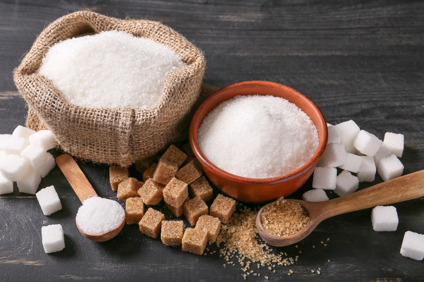 Different types of sugar, white and brown, in granules and cubes, in a sack, bowl, and two spoons on a table.