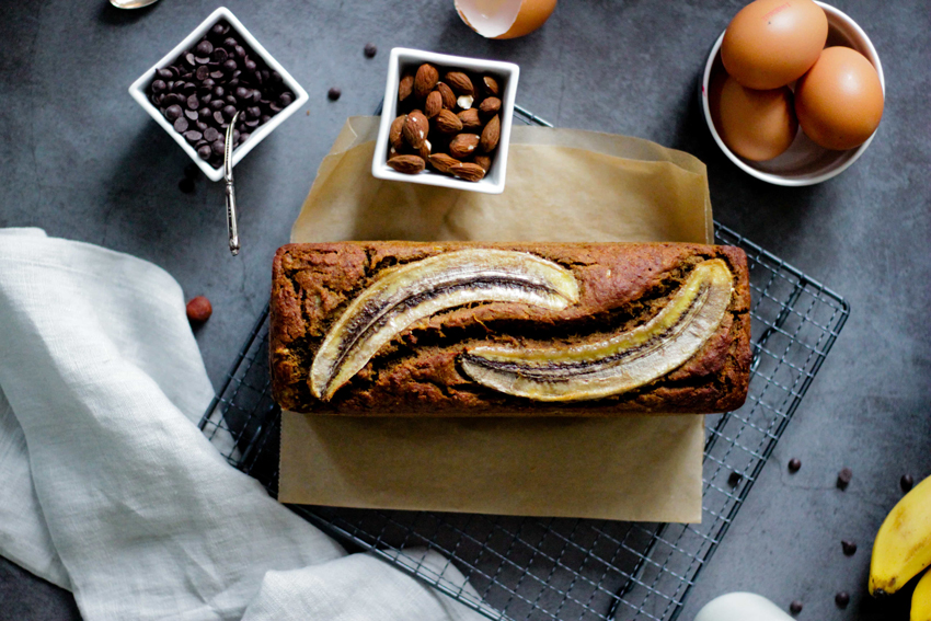 Freshly baked banana bread on a table with baking tray, parchment paper, a cloth, and dessert ingredients like eggs, nuts, chocolate chips, and bananas.