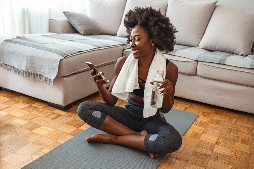 Woman in sportswear sitting cross-legged on a mat at home, smiling at her phone, holding a water bottle.