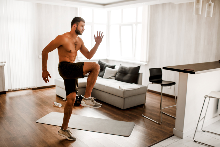 Fit shirtless man doing a Tabata exercise standing in his living room with a mat and water bottle nearby.